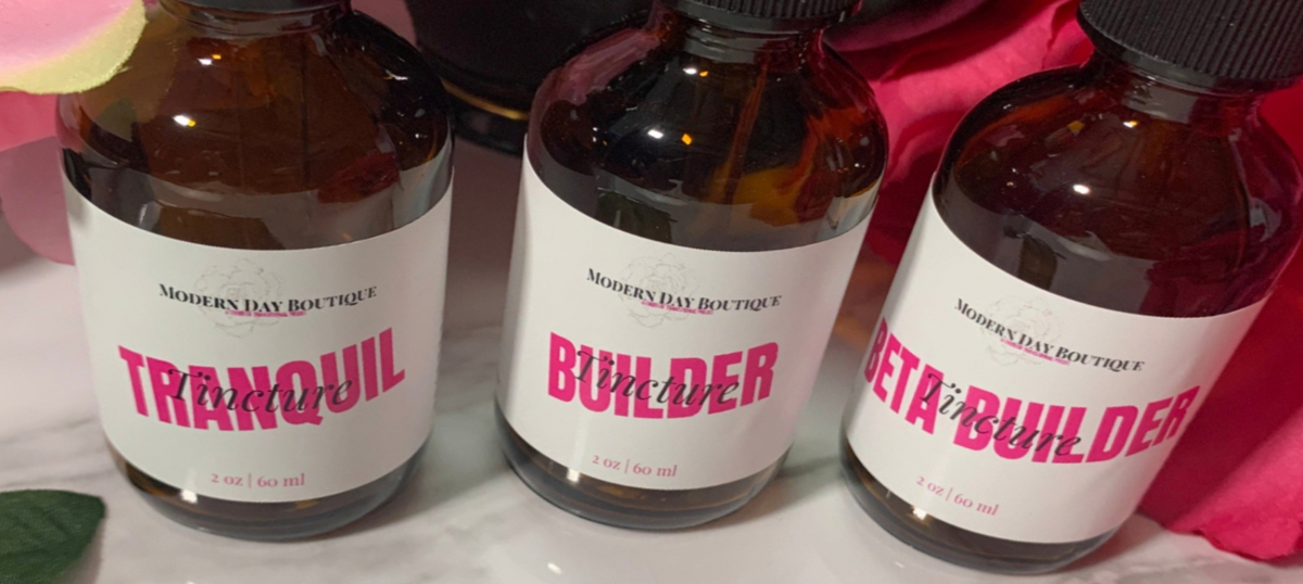 Three amber glass bottles with white labels from Modern Day Boutique, labeled Tranquil, Builder, and Beta Builder, arranged on a white surface with pink fabric in the background.